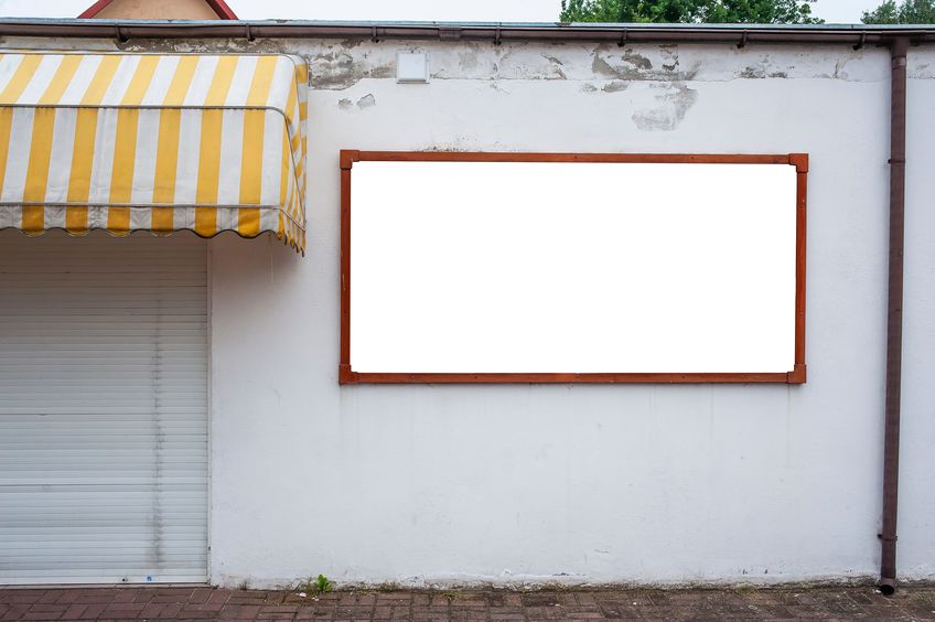 Pannello per insegna vuoto con cornice in legno su un muro bianco, accanto a una tenda da sole a righe gialle