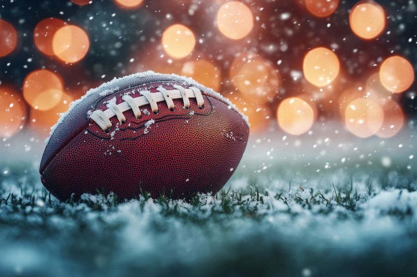 Pallone da football americano coperto di neve su un campo innevato, con luci sfocate sullo sfondo.