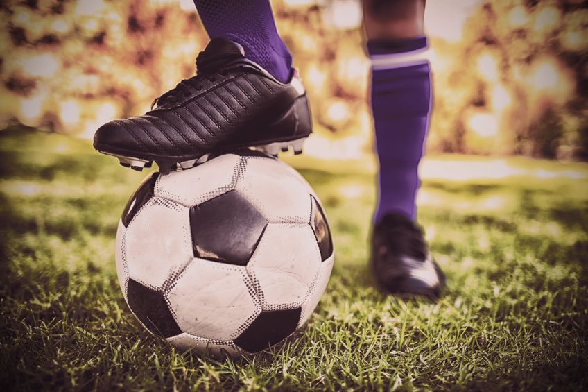 Piede con scarpino da calcio nero appoggiato su un pallone, su un prato, con effetto fotografico vintage.