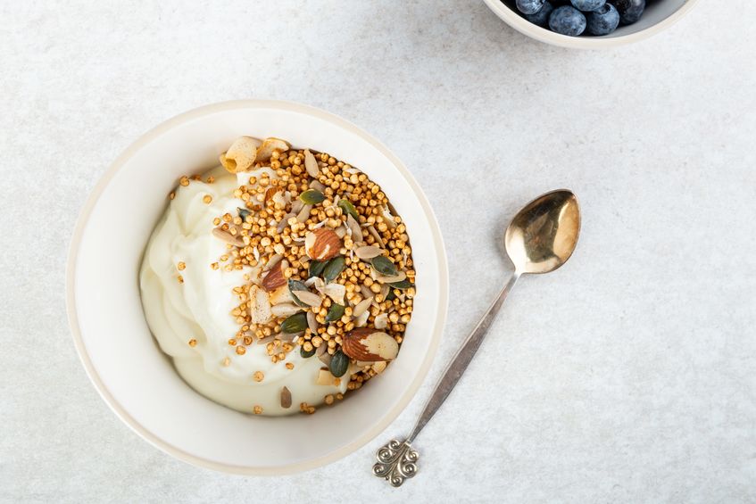 Coppetta di yogurt bianco con cereali soffiati, mandorle e semi, vista dall'alto, accanto a un cucchiaino.